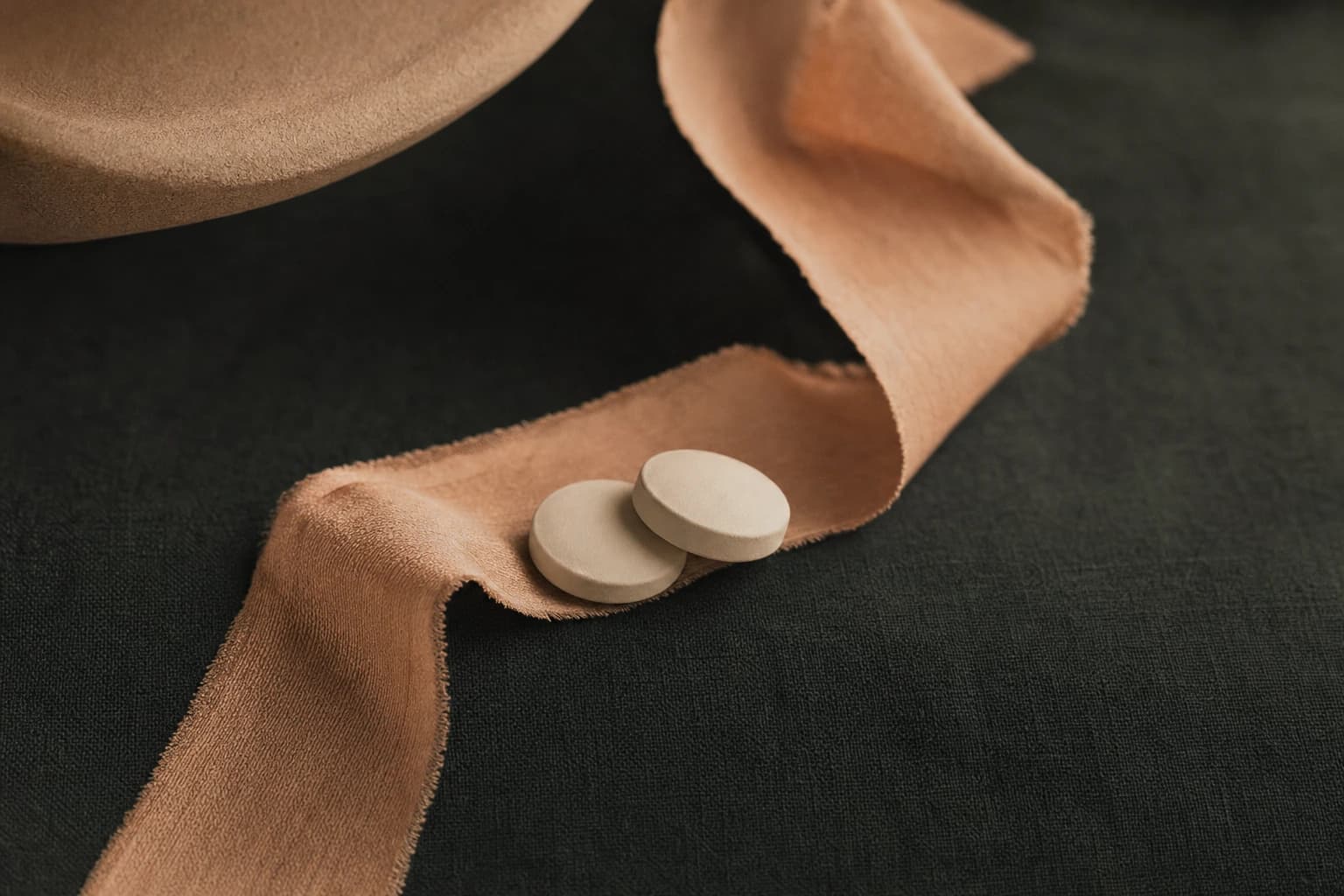 Detailaufnahme — bone-farbene Tabletten und ein warmes Seidenband auf anthrazitfarbenem Leinen