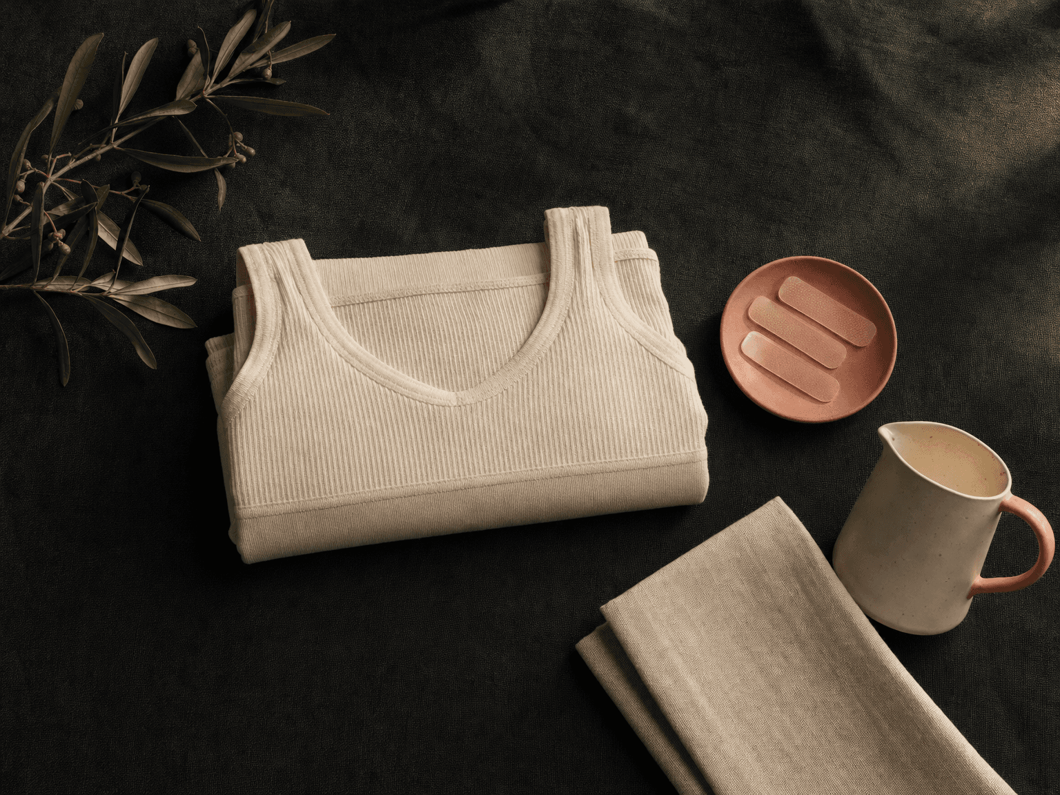 Gefalteter bone-cream Stütz-BH ohne Front-Zip, kleine Tonschale mit Silikon-Scar-Pflastern, Olivenzweig und Linentuch auf charcoal-grünem Leinen — Stillleben Nachsorge