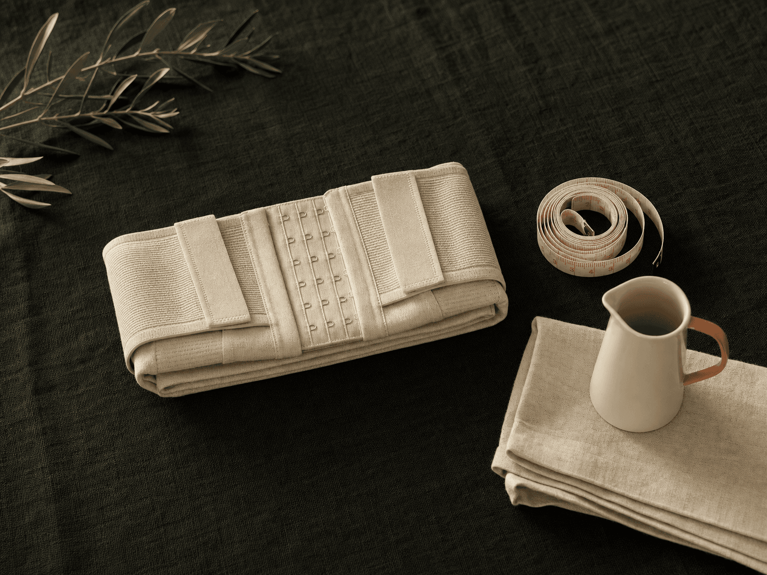 Gefalteter bone-cream Heavy-Support-BH mit Hook-Eye-Front und Velcro-Riemen, daneben aufgerolltes Maßband, Olivenzweig und Linentuch auf charcoal-grünem Leinen — Stillleben Nachsorge