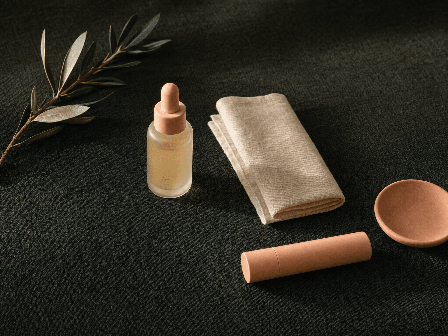 Apothecary glass bottle of antiviral medication, a slim peach lip-balm case, a linen cloth and an olive branch on charcoal-green linen — an aftercare still life
