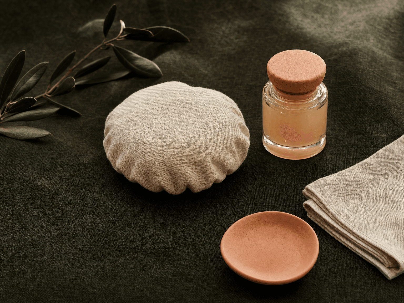 Kleiner bone-cream Kühl-Pad, Apotheker-Glas mit Arnika-Gel, Olivenzweig und Linentuch auf charcoal-grünem Leinen — minimalistische Nachsorge