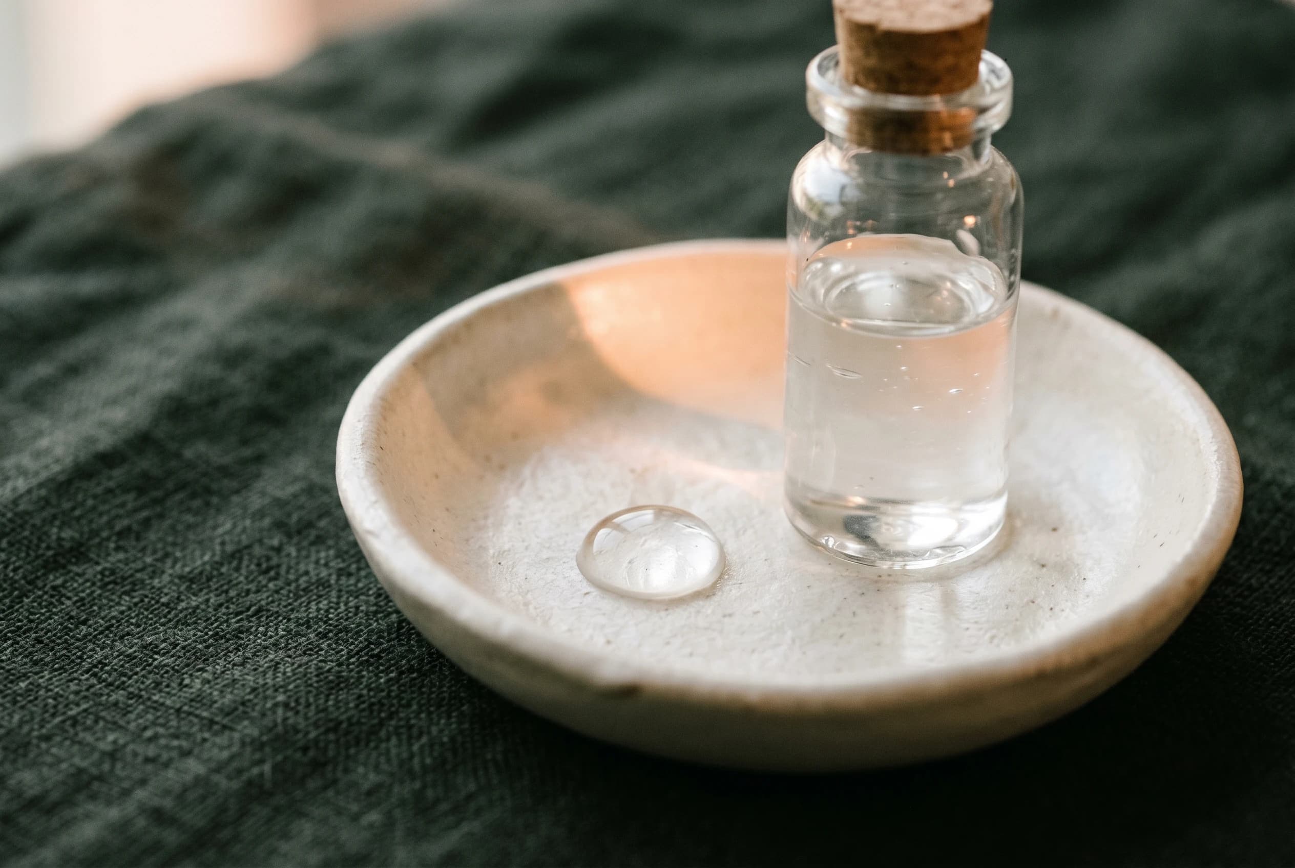 Clear hyaluronic acid gel in a small vial and a droplet in a bone-cream ceramic dish on charcoal-green linen — a symbol of reversible hyaluronic acid