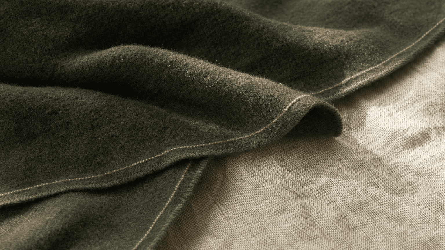 A charcoal-forest wool blanket with a peach hairline trim on bone-cream linen — calm, warm recovery texture