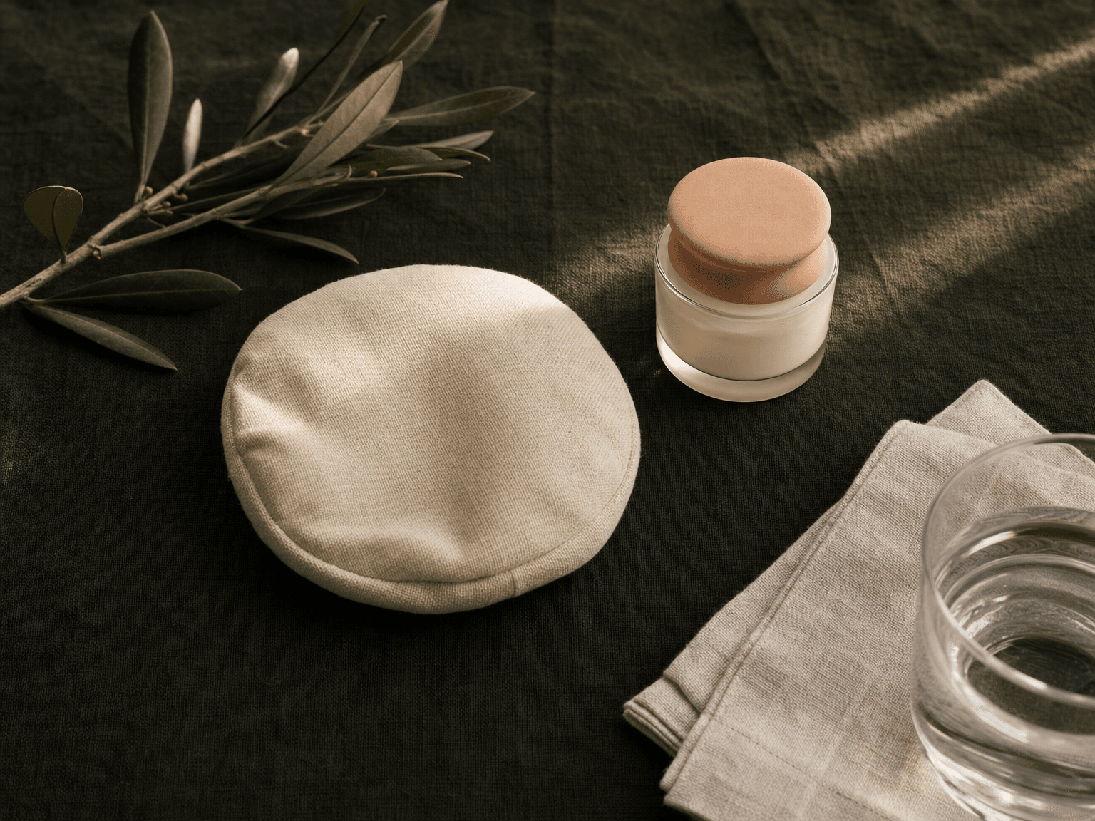 Stilllife: kleines bone-cream Kühl-Pad, Apotheker-Glas mit Pflegecreme, Mineralwasser und Linentuch auf charcoal-grünem Leinen