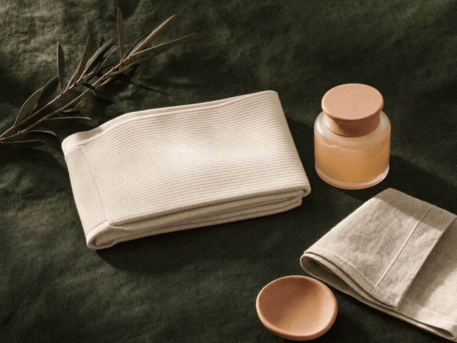 Gefaltete bone-cream Kompressionswäsche mit Arnika-Glas und Olivenzweig auf charcoal-grünem Leinen — Stillleben Nachsorge
